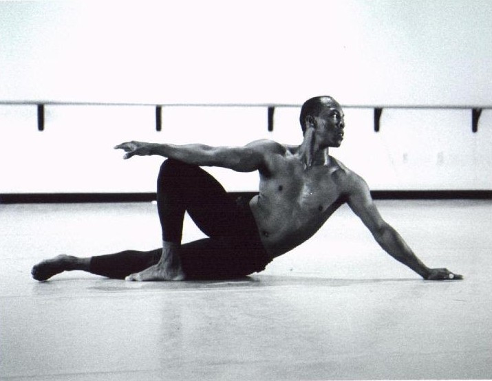 Black-and-white photograph of Boris Willis seated on the studio floor, one arm extended, beside a ballet barre