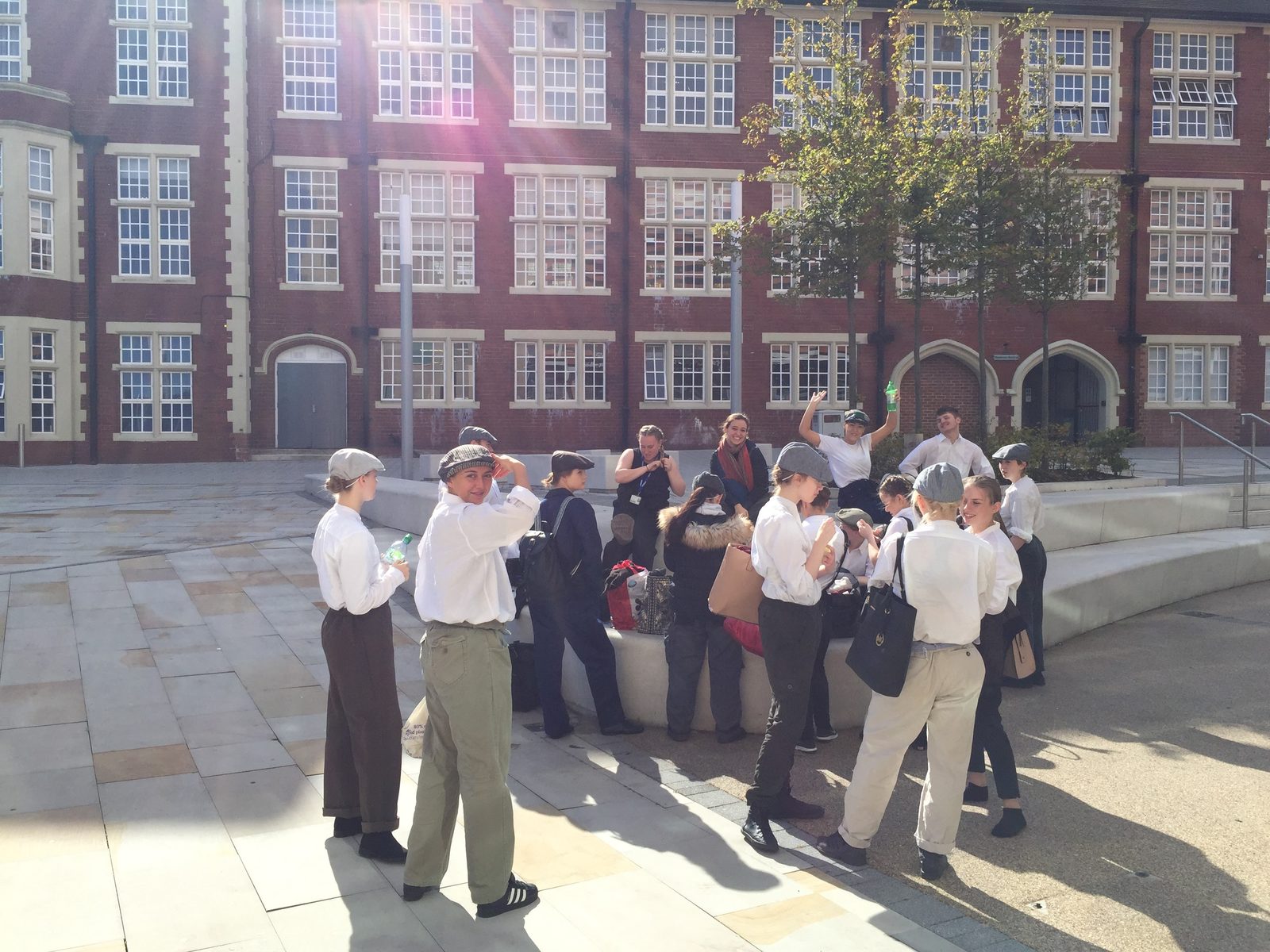 Dancers in period workwear and flat caps gathered outside a red-brick building